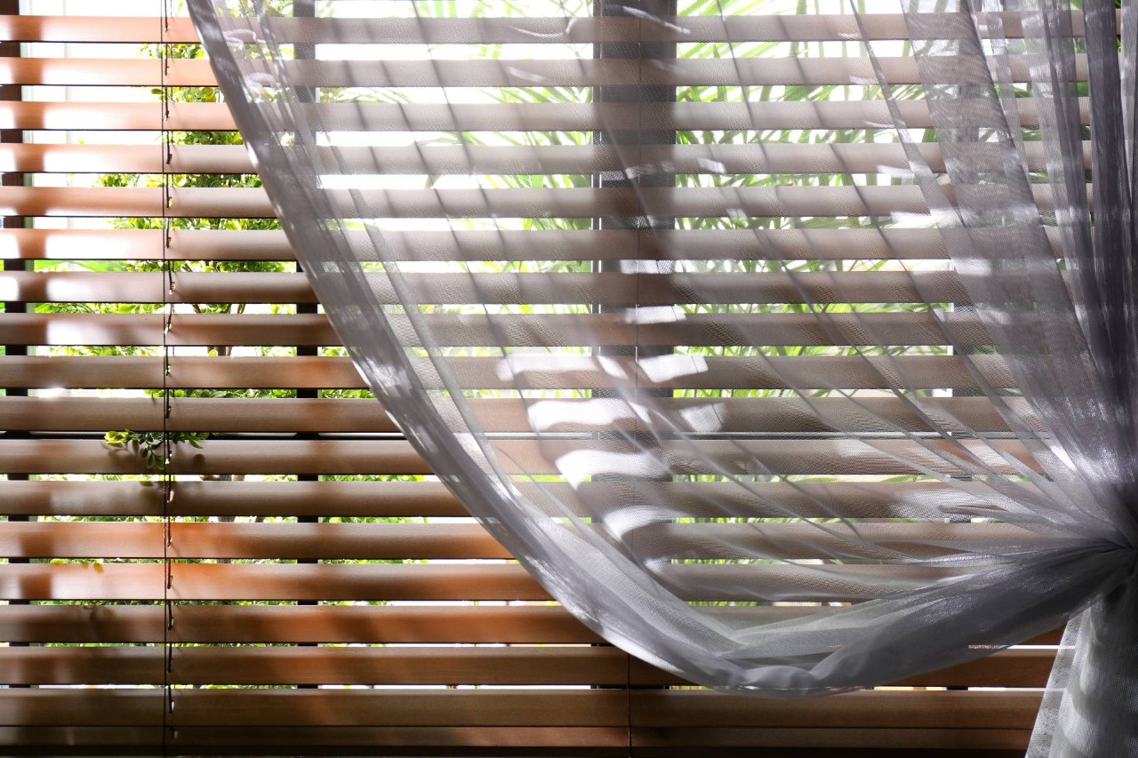 Layered window treatments, wood blinds with sheer drapes, on a home's interior window with greenery outside at Hubbard's Custom Blinds near Cambridge, Maryland (MD)